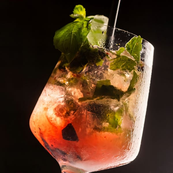 Close-up of a vibrant red cocktail with ice, mint, and basil in a condensation-covered glass.