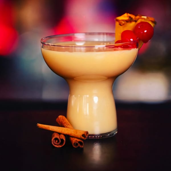 A creamy Haitian cocktail garnished with pineapple and cherries, with cinnamon sticks on a dark surface.