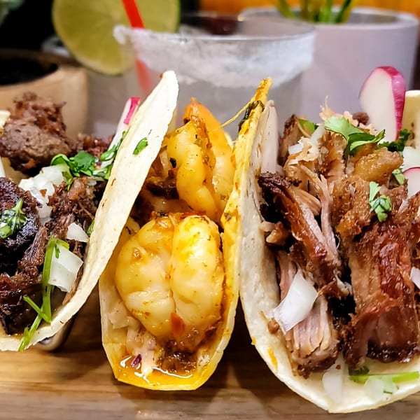 Three Haitian tacos, one with beef, one with shrimp, and one with pork, on a wooden board.